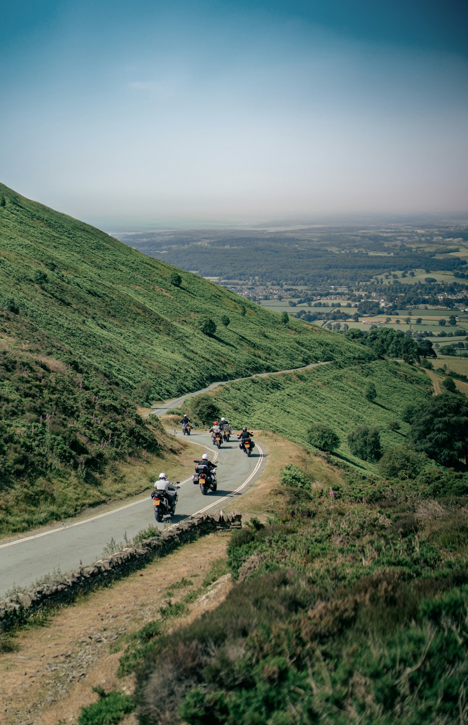 Motorcyclists ride down a winding road on a green hillside.