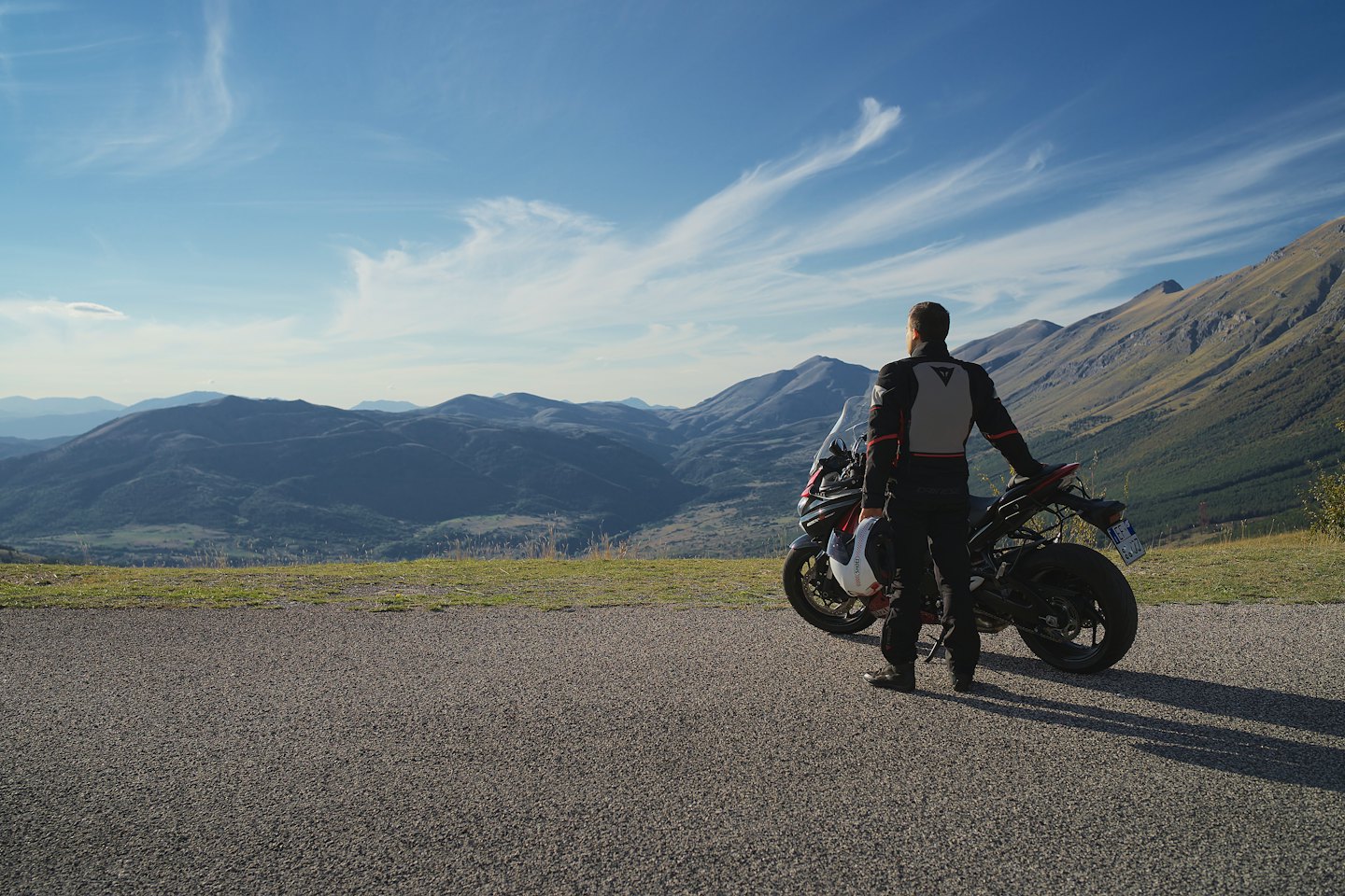 Motorcyclist with helmet stands by motorcycle overlooking mountains.